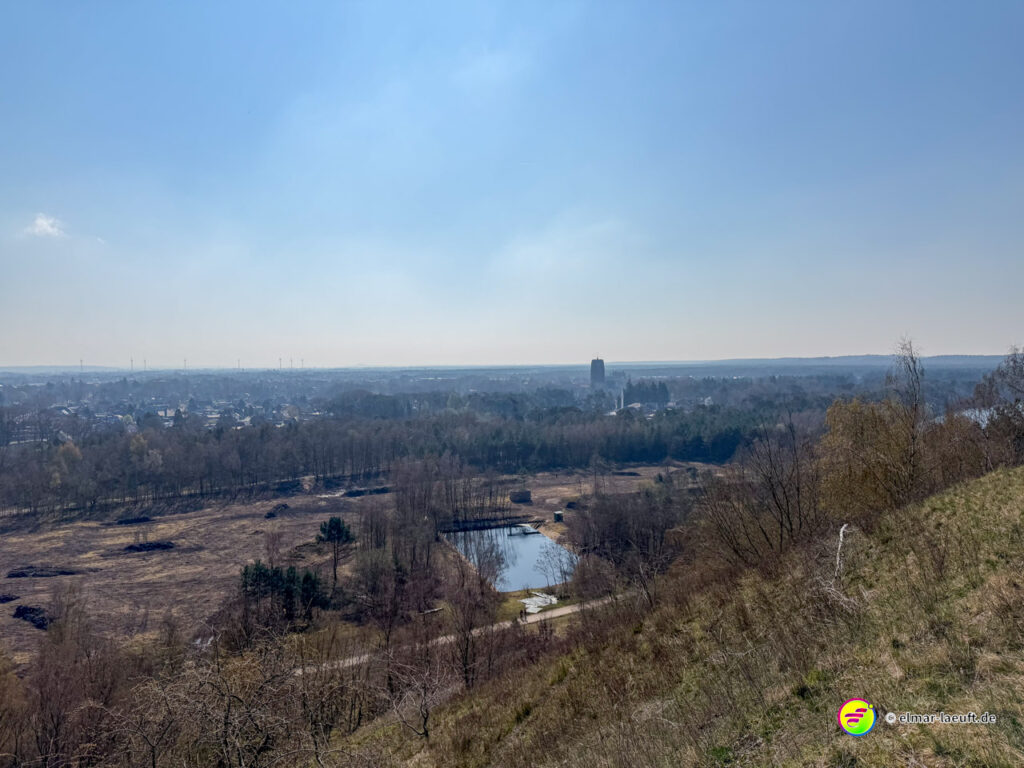 Blick beim Laufen in Maasmechelen von einem Hang auf eine offene Landschaft mit Bäumen, einem kleinen Teich und einer Stadt im Hintergrund.