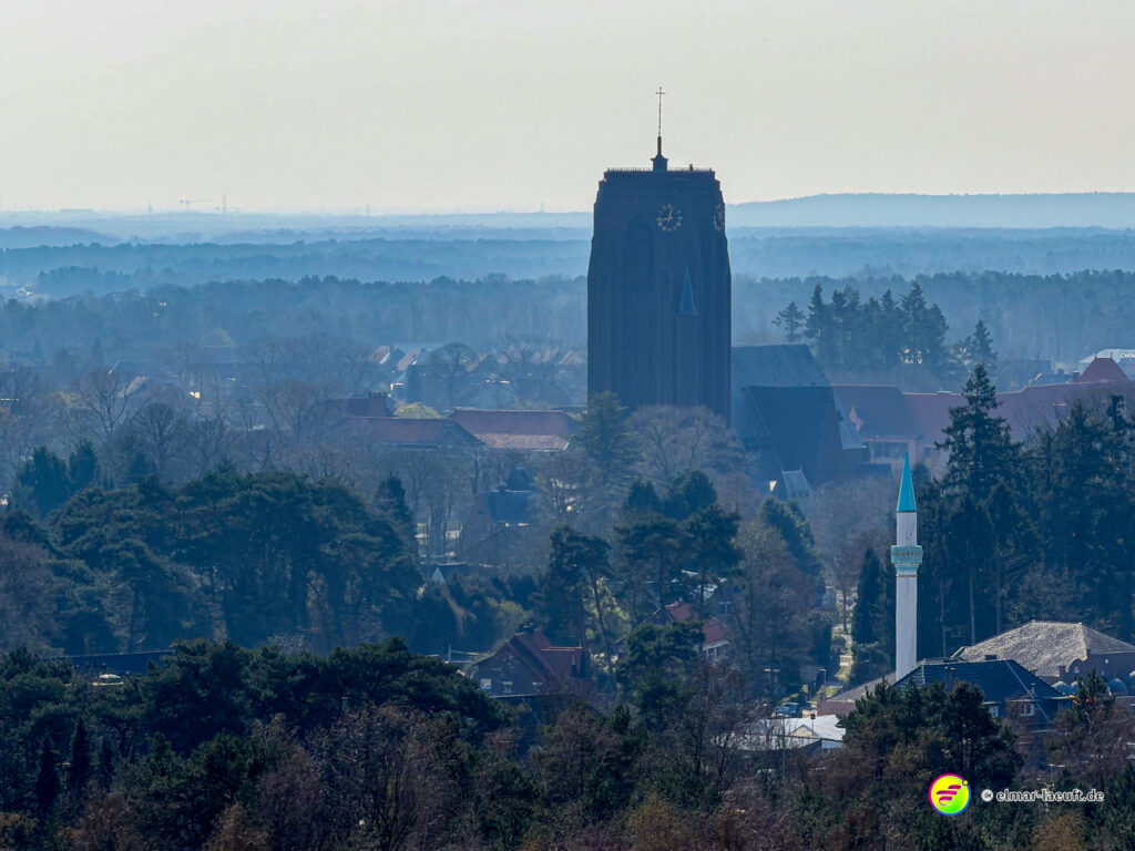 Blick beim Laufen auf Maasmechelen mit einer markanten Kirche und viel Wald im Vordergrund.
