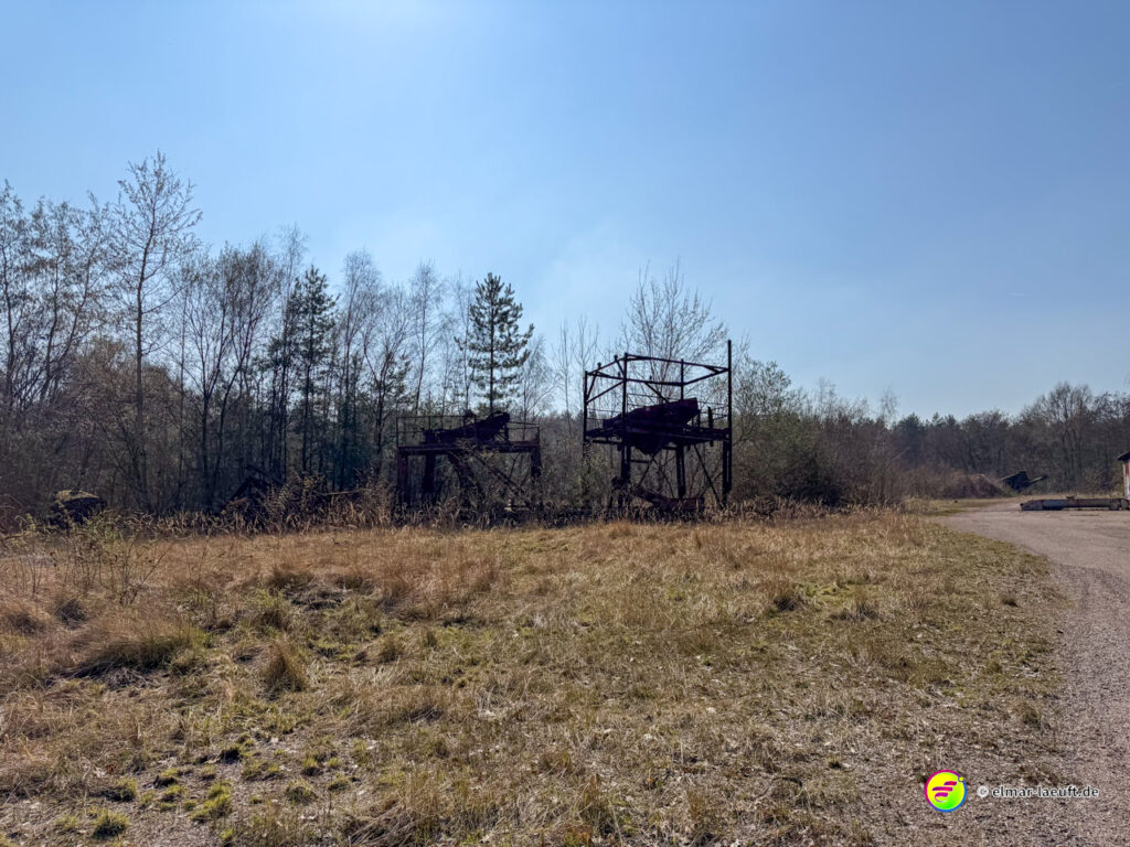 Laufen auf einem abgelegenen Weg in Maasmechelen mit verwildertem Gras und alten, rostigen Industrieanlagen am Waldrand unter klarem Himmel.