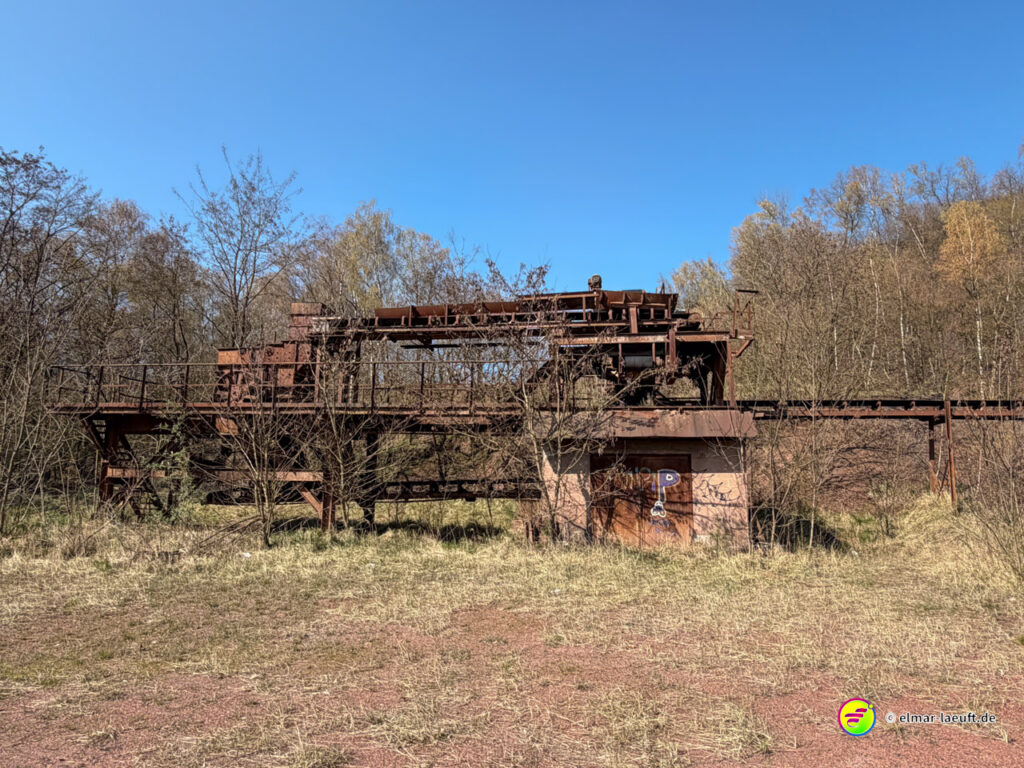 Laufen entlang einer verlassenen, rostigen Industrieanlage in einer offenen, von Bäumen umgebenen Landschaft bei klarem Himmel.