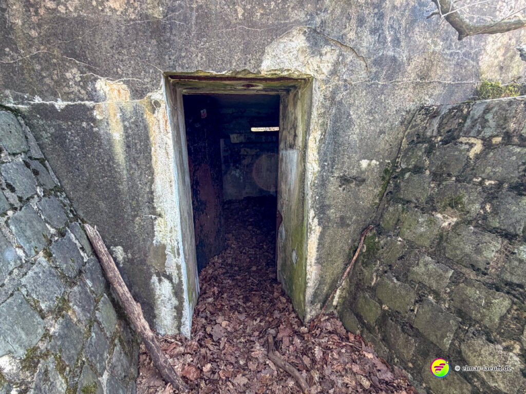 Laufen auf einer Entdeckungstour in Maasmechelen führt an einem alten, moosbewachsenen Bunker mit einem dunklen Eingang vorbei, umgeben von herbstlichen Blättern.