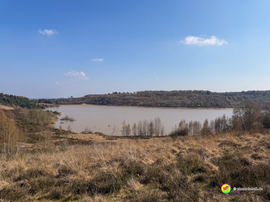 Laufen in der offenen Natur bei Maasmechelen mit Blick auf einen See und hügelige, teils bewaldete Landschaft unter klarem Himmel.