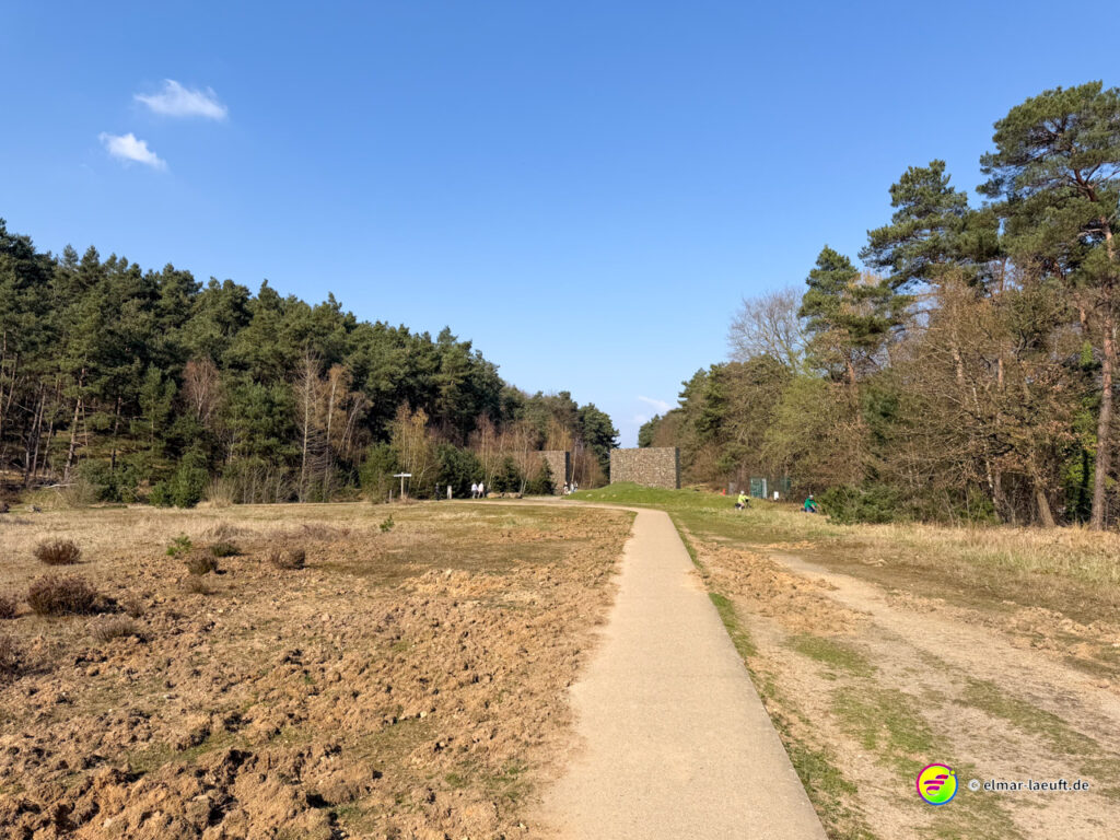 Laufen auf einem schmalen, befestigten Weg durch eine offene Landschaft mit Wald am Rand und einer markanten Steinmauer im Hintergrund bei klarem Himmel in Maasmechelen.