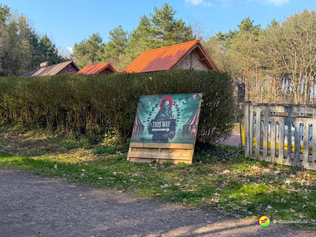 Laufen auf einem Waldweg in Maasmechelen mit einem auffälligen Schild eines Clowns, das den Weg weist, vor einer Hecke und kleinen Häusern mit roten Dächern.