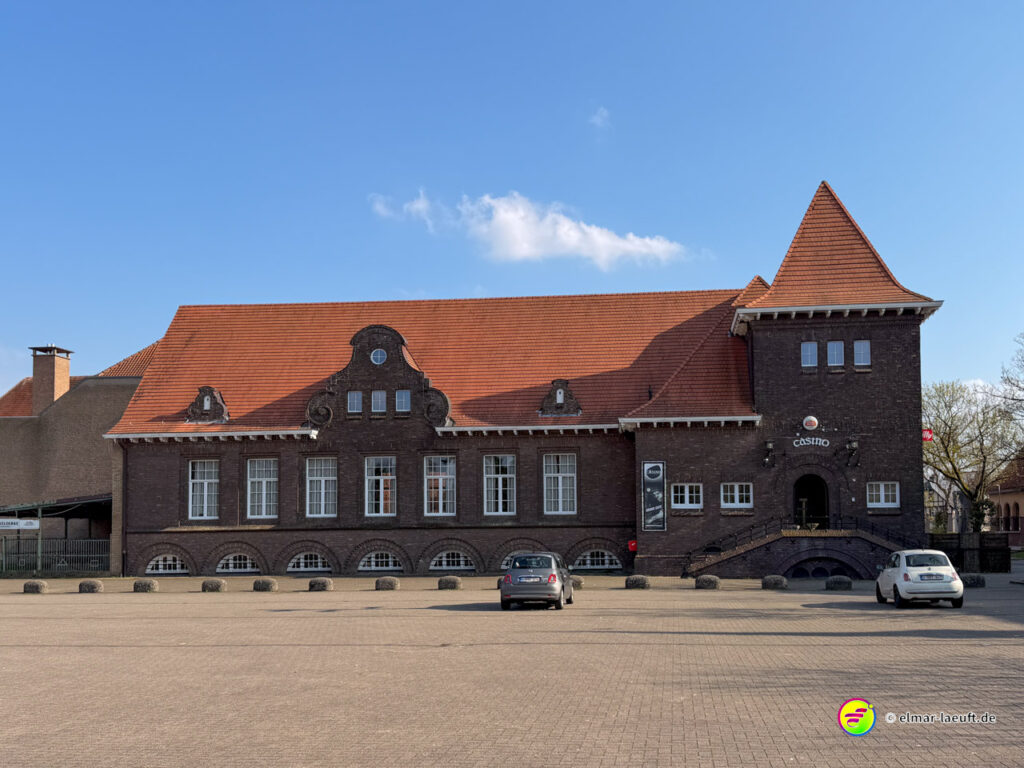 Laufen in Maasmechelen mit Blick auf ein historisches Gebäude mit rotem Ziegeldach und kleinem Parkplatz im Vordergrund.