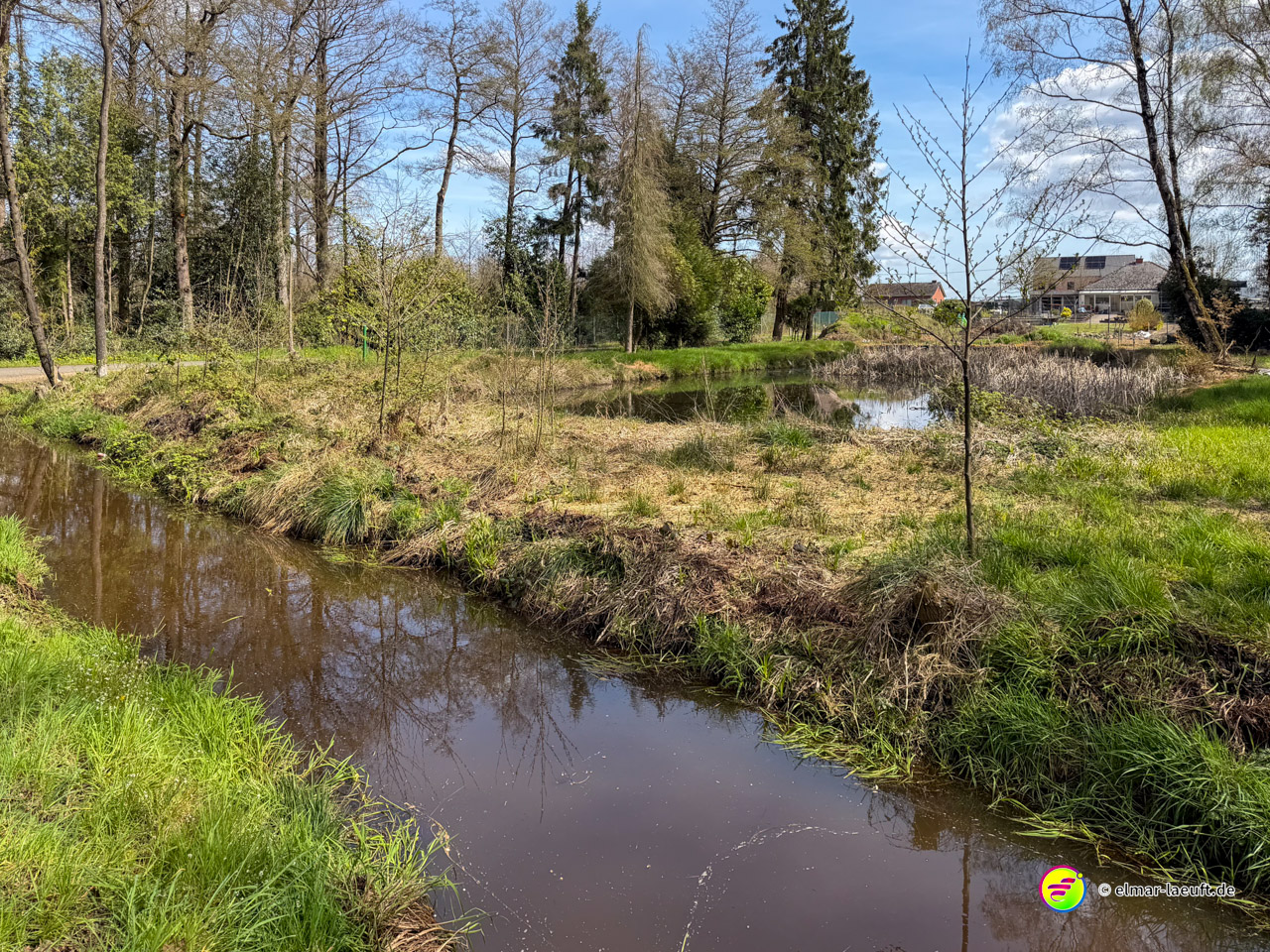 Laufen entlang eines ruhigen Baches mit grünen Uferbereichen und Bäumen in Oplagbeek, mit Häusern im Hintergrund.